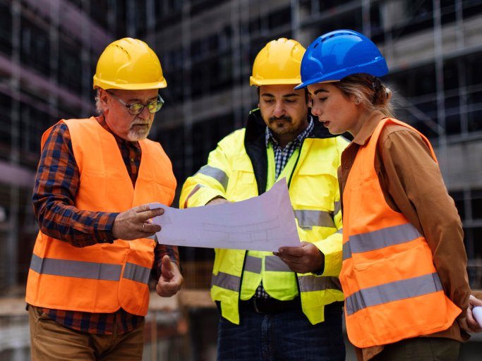 Three construction site managers wearing safety gear, looking over a blueprint