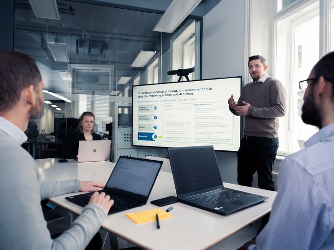 Three people sitting with their laptops at a table while another person stands against a large screen while presenting to them