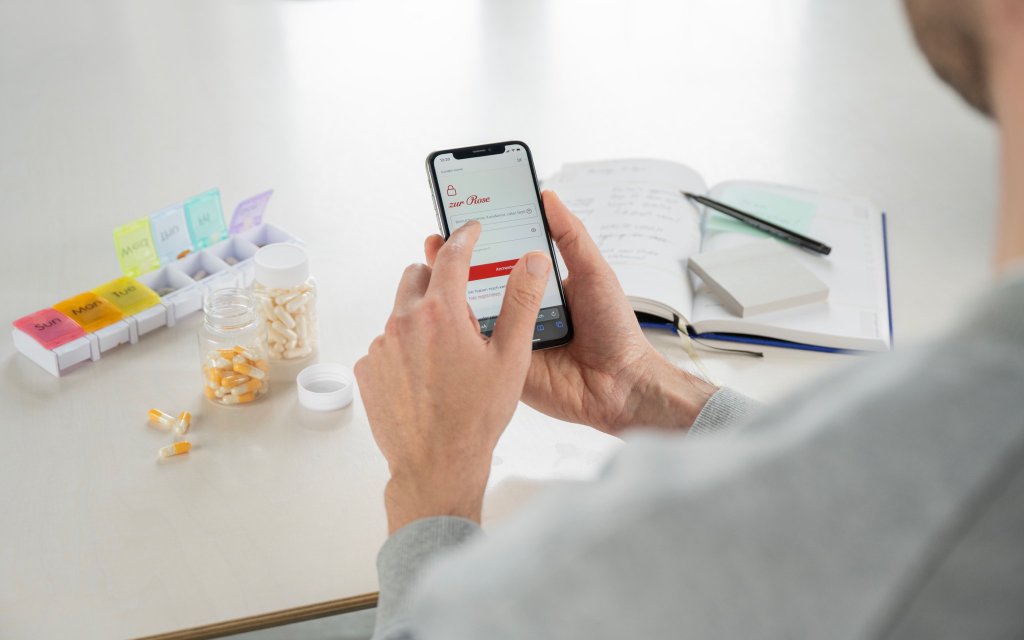 Hände halten ein Mobiltelefon. Im Hintergrund befinden sich Pillen, ein Pillen-Organizer und ein Notizbuch