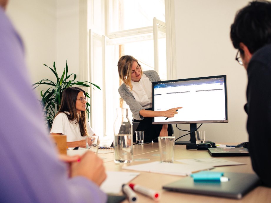 3 Personen sitzen während eines Workshops an einem Tisch, während eine Frau etwas präsentiert und auf einen Bildschirm zeigt