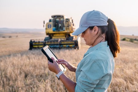 Moderne Bäuerin in einem Weizenfeld, die ein IoT-Tablet überprüft, mit einem Weizenhäcksler im Hintergrund